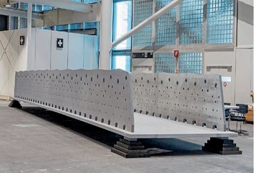 Das Bild zeigt ein großes, vorgefertigtes Brückenelement aus Beton, das in einer modernen Halle auf mehreren schwarzen Unterlagen gelagert ist. Die Brückenplatte besitzt seitliche, gelochte Wände und wirkt massiv sowie technisch präzise gefertigt. Im Hintergrund sind Glasbausteine, große Fenster und industrielle Ausstattung zu erkennen, was auf eine Fertigungshalle oder ein Prüflabor hinweist.

                                  